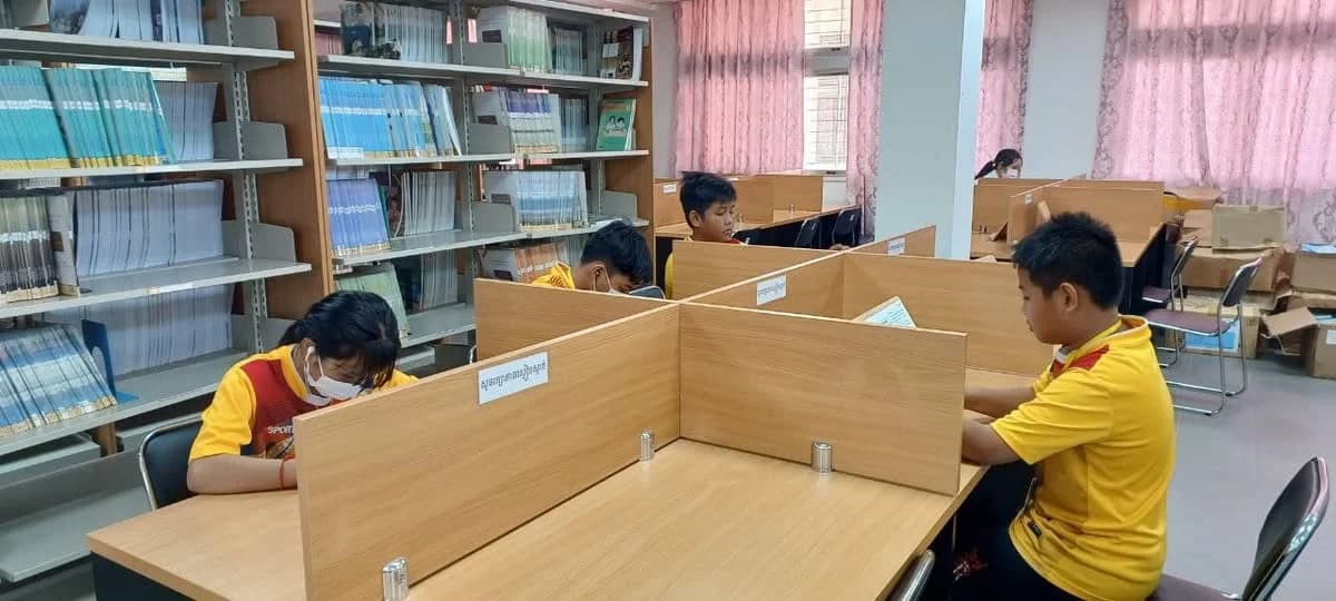 Students sitting at individual study carrels with partitions, reading books and studying in a library with bookshelves in the background.