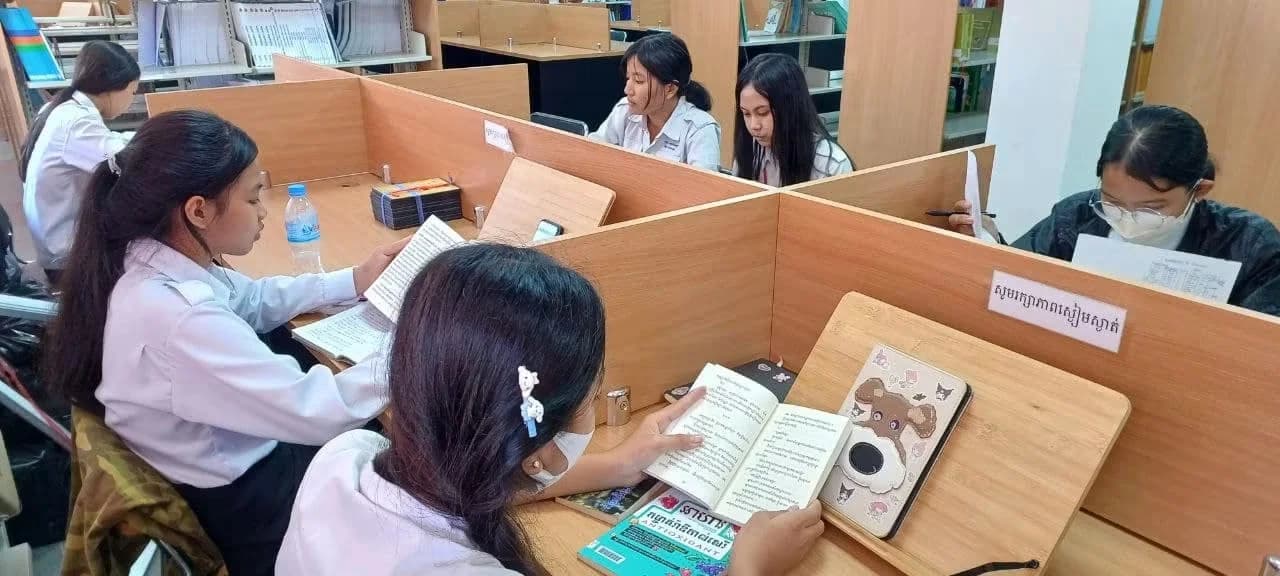 Students in school uniforms sitting at individual partitioned study desks, reading books and studying in a library.