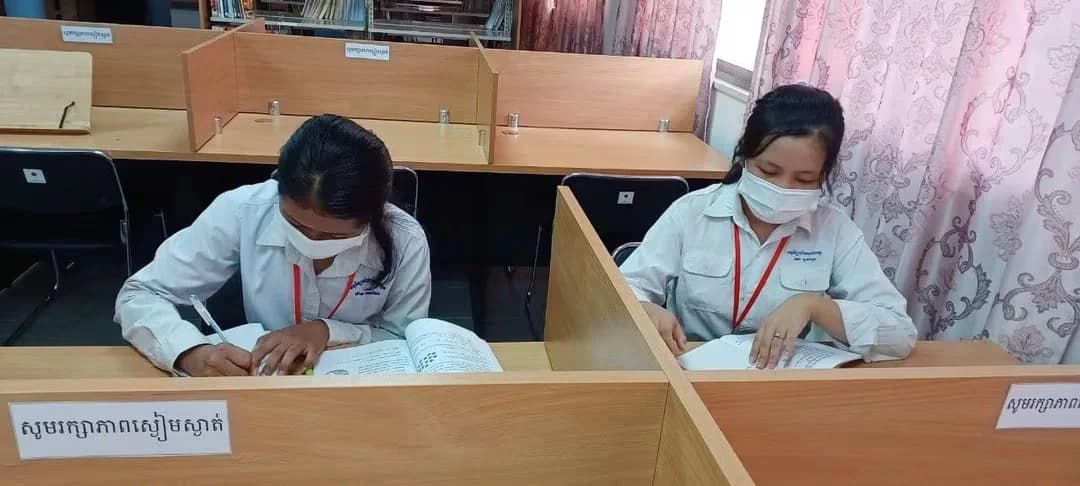 Two female students wearing face masks, sitting at partitioned study desks writing notes and reading documents in a library.